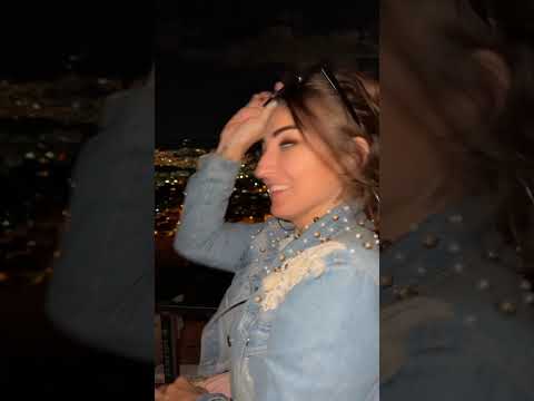 A woman with long hair and sunglasses plays with her hair while enjoying a nighttime cityscape in the background.