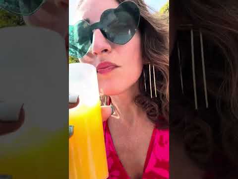 A woman in oversized heart-shaped sunglasses sips a vibrant orange drink, adorned in a bright pink top, enjoying a sunny day outdoors.